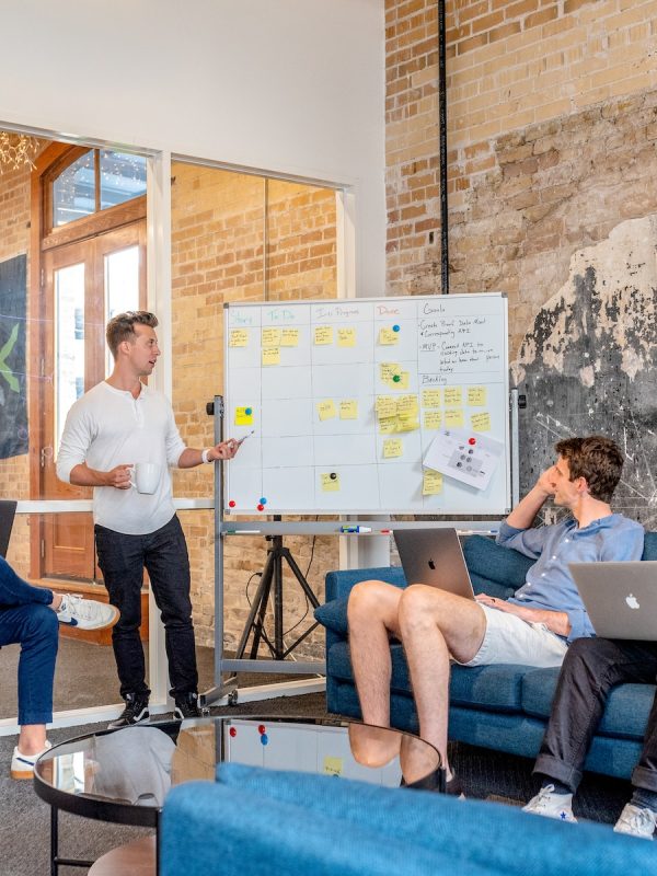 three men sitting while using laptops and watching man beside whiteboard