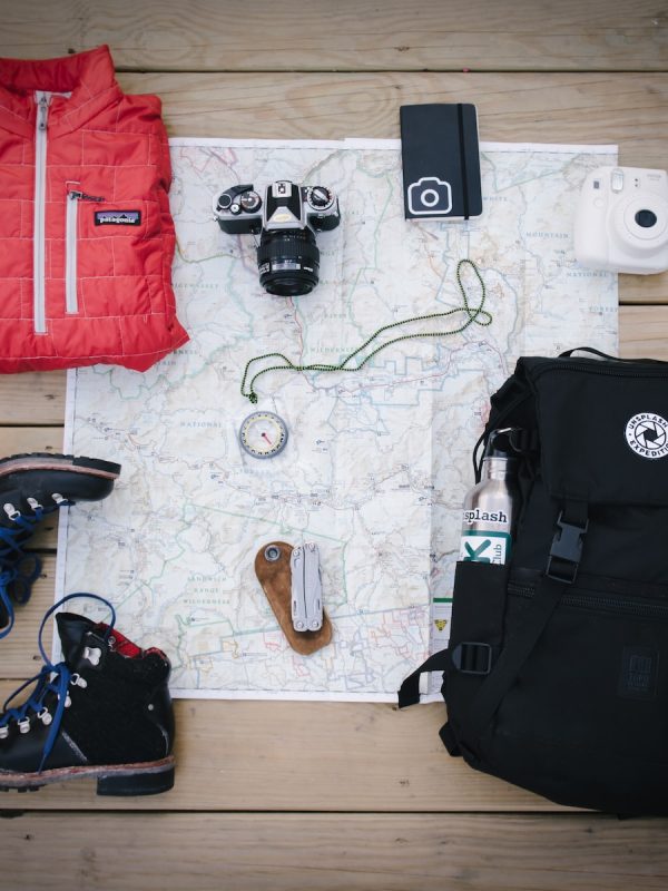 black hiking backpack near white Fujifilm instax mini camera near black leather boots, red half-zip jacket, gray pocket watch on white map